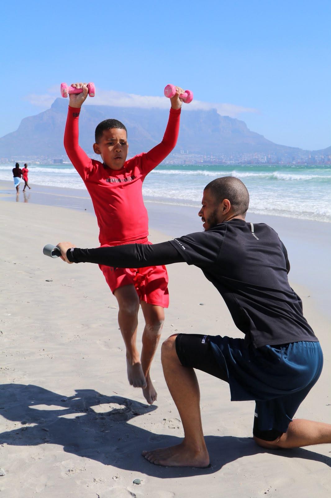 Athlete pressing dumbbells on the beach with a coach